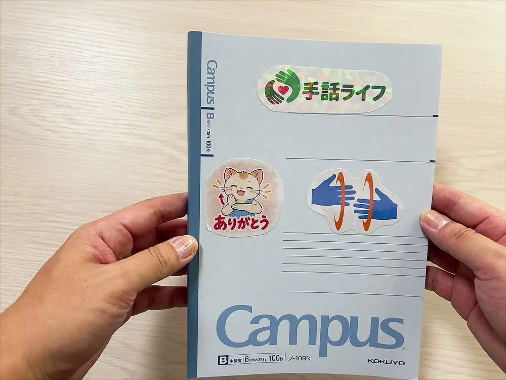 手話ライフのロゴと猫のキャラクター、手のイラストのホログラムシールを貼ったノートの表紙