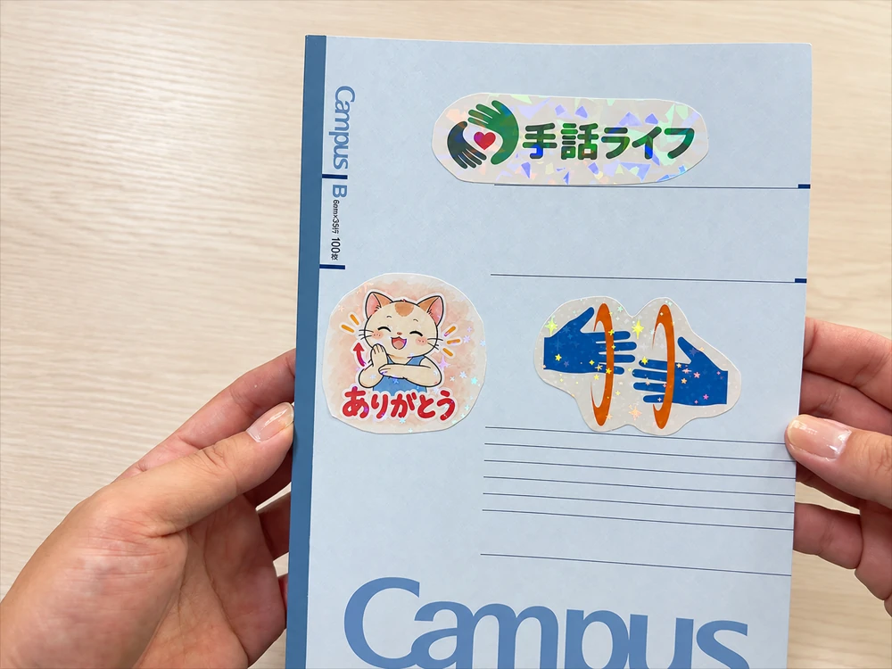 完成したノートの表紙のアップ。手話ライフのロゴや猫のキャラクター、手のイラストのシールに、虹色や星のホログラムが光っている