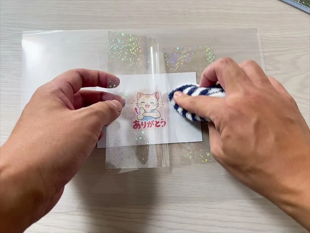 印刷した猫のキャラクターに透明なホログラムシートを重ね、クリアファイル越しに布で押さえている手元