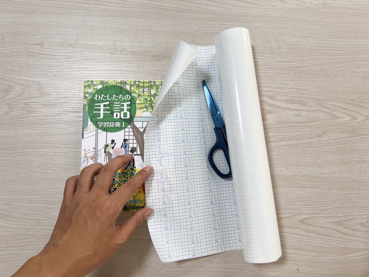 『わたしたちの手話 学習辞典Ⅰ』と書籍用保護フィルム、ハサミを準備した様子